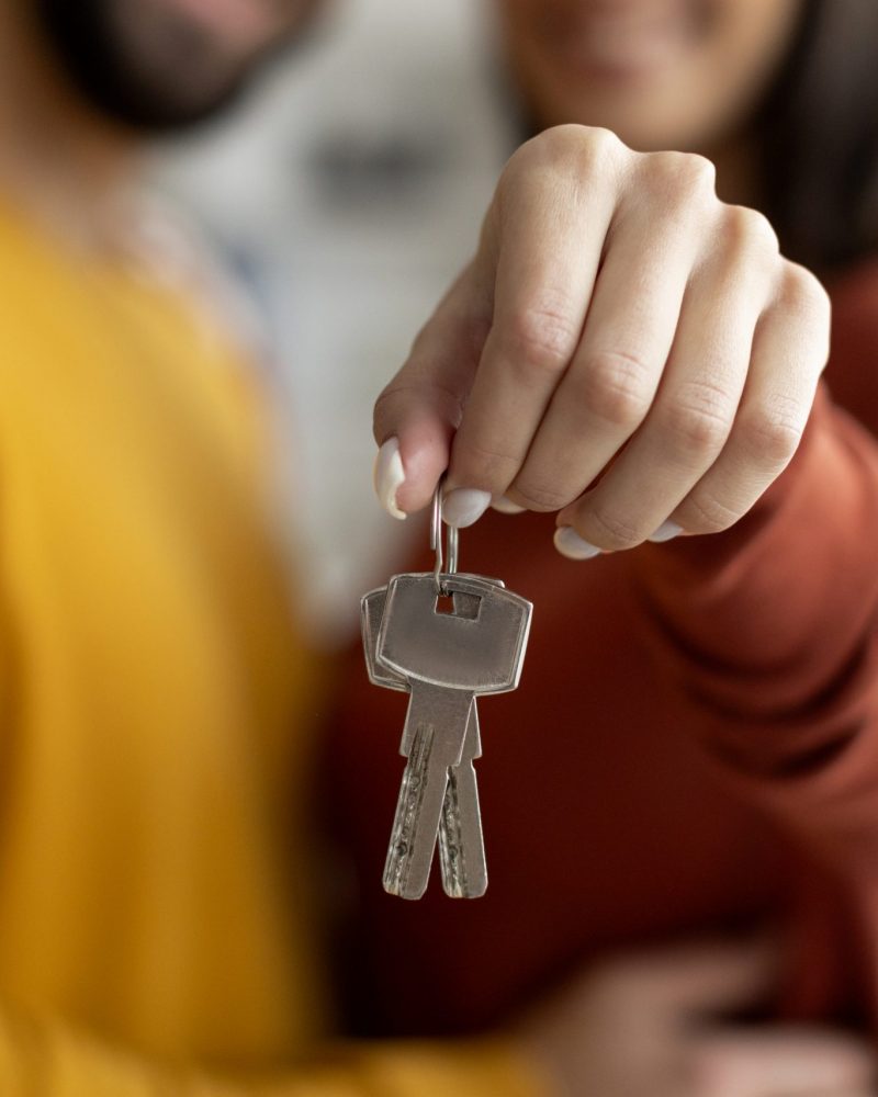 Closeup Shot Of Romantic Young Couple Holding Home Keys In Hands, Loving Millennial Spouses Celebrating Moving To Their Own House, Embracing And Smiling, Cropped Image With Selective Focus