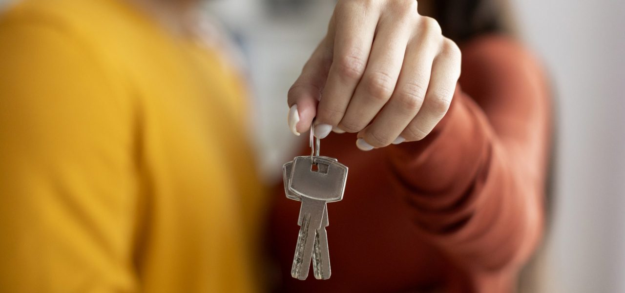 Closeup Shot Of Romantic Young Couple Holding Home Keys In Hands, Loving Millennial Spouses Celebrating Moving To Their Own House, Embracing And Smiling, Cropped Image With Selective Focus